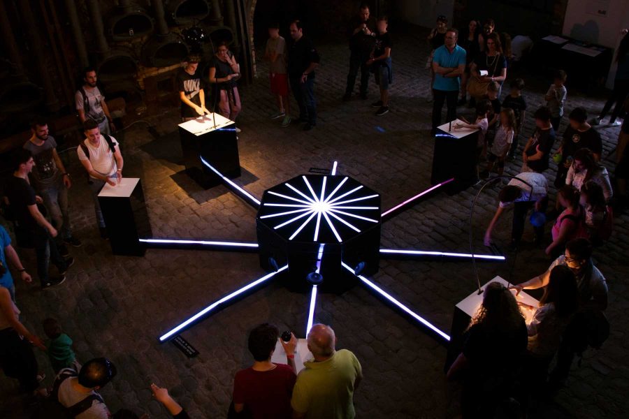 Participants play instruments around the illuminated central hub in a dark exhibition space.